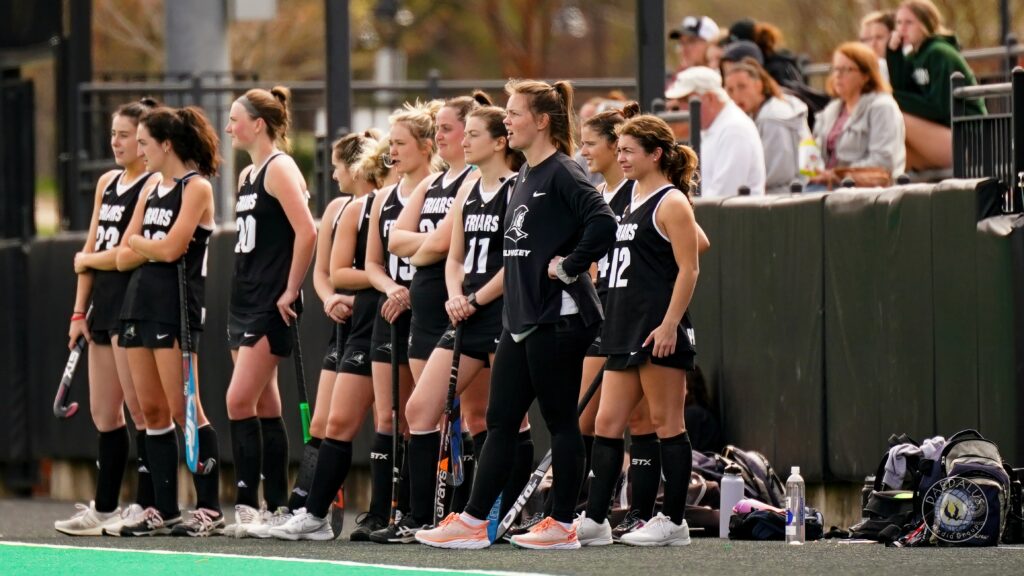 field hockey players in black uniforms on sideline during a game